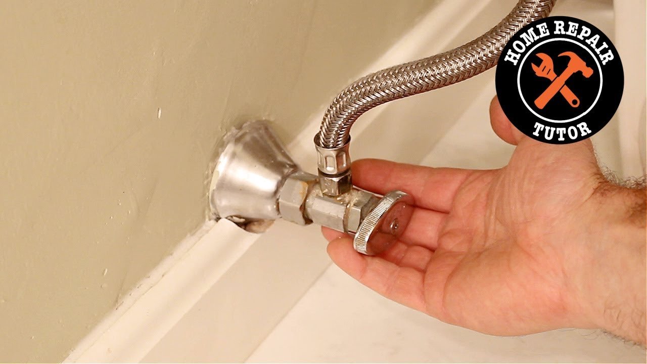 A person reaches down to check the toilet shut-off valve in a bathroom.